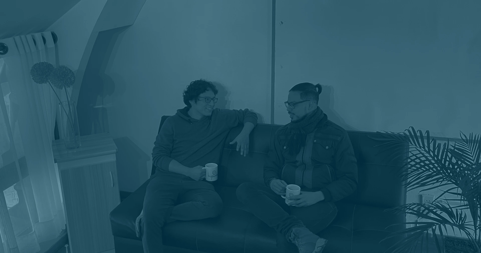 A group of men sitting on a couch indoors, captured in black and white. They are dressed in various clothing styles, with visible human faces. The setting includes a wall behind them