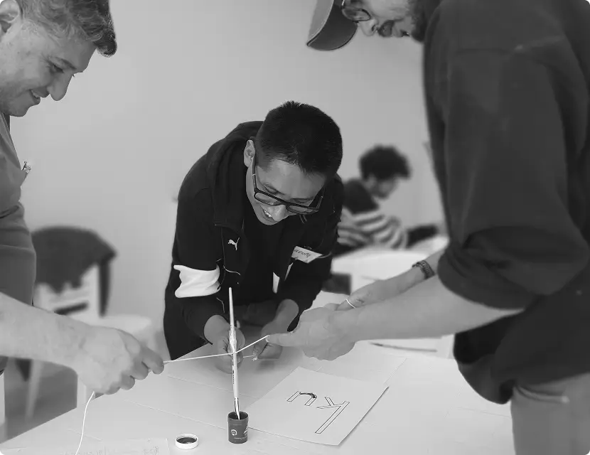 A group of diverse individuals collaborating on a project at a table in an indoor setting, surrounded by office supplies and artwork on the walls. The scene conveys focus and teamwork