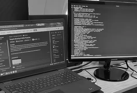 A laptop and a monitor are positioned on a desk, surrounded by office supplies. The display shows some text content, while the laptop sits closed next to the monitor. Both devices represent typical computer hardware used in an indoor office environment