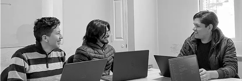 A group of people sitting at a table in an indoor office setting, each using laptops. There is a window in the background, and the individuals are engaged in their work