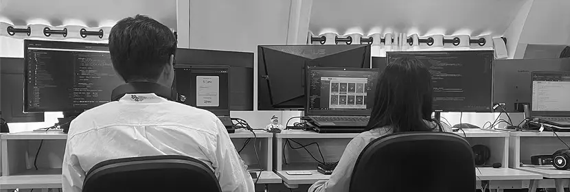 A man and a woman sitting at desks in an office building, focused on their computers. The environment includes computer monitors, chairs, and a wall in the background. The scene is captured in black and white.